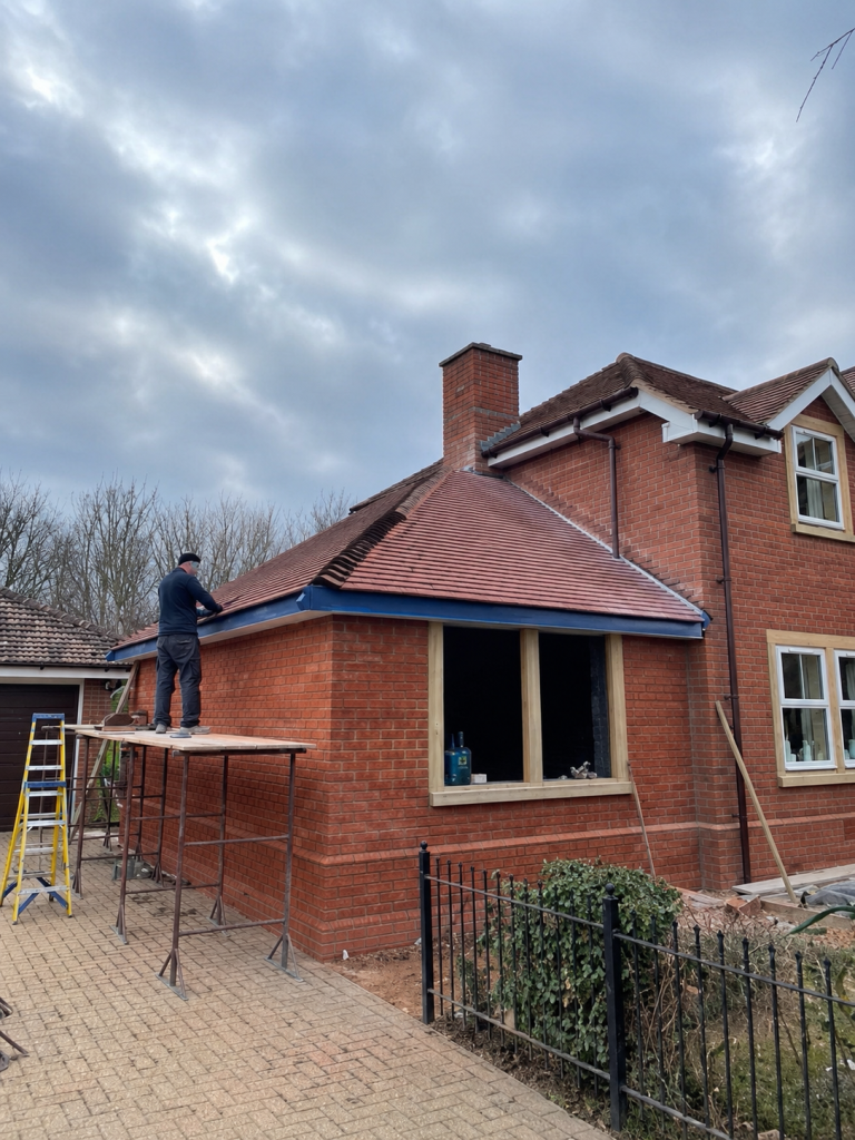 Builder working from scaffolding on the tiled roof of a red-brick house extension
